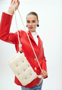 Borsa a tracolla in pelle bianca con dettagli in oro e un nodo sulla tracolla, tenuta da una persona che indossa un blazer in pelle rossa.