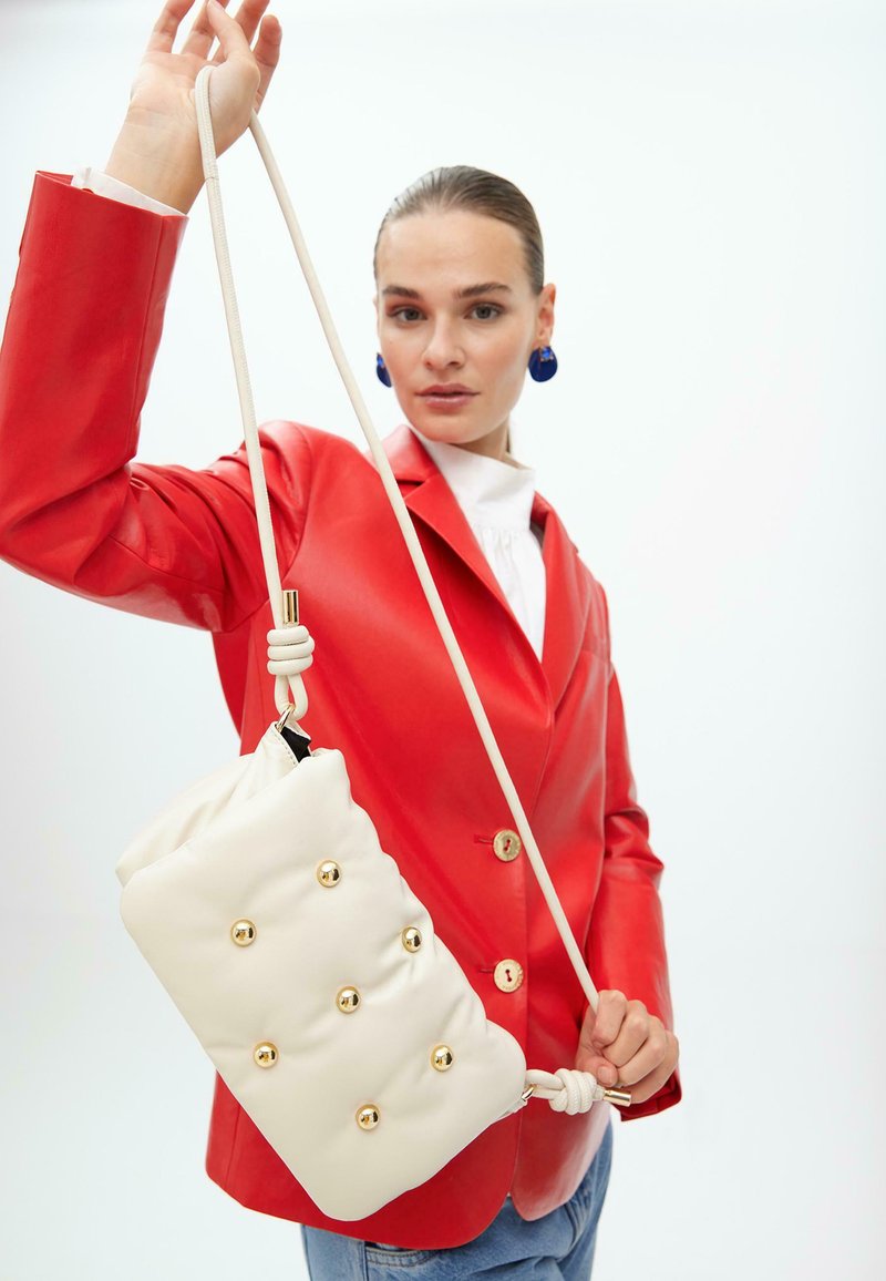 Borsa a tracolla in pelle bianca con dettagli in oro e un nodo sulla tracolla, tenuta da una persona che indossa un blazer in pelle rossa.