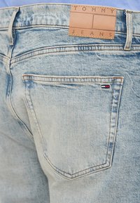 Svetlo modré jeany z denimu s koženou nášivkou označenou "TOMMY JEANS" na páse. Obsahujú švy a malé logo na zadnej vrecku.