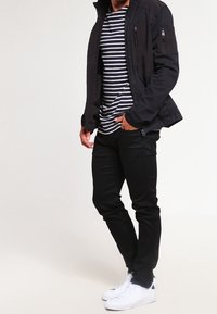 Veste noire avec des zippers, chemise rayée noir et blanc, pantalon noir et baskets blanches. Textures douces avec un design décontracté.