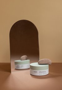 Barattolo rotondo con un coperchio verde menta e una base bianca, contenente patch per occhi al Caffè e Tè Verde, sormontato da una pietra marrone liscia, che si riflette in uno specchio.