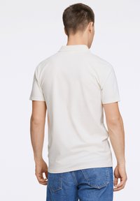 Krämfärgad polo-tröja med klassisk krage, korta ärmar och rak nederkant, i kombination med blå jeans. Enkel, odekorerad design.