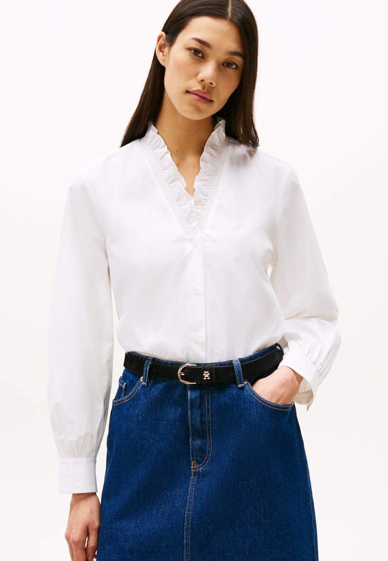 Blusa de algodón blanca con un escote con volantes, mangas largas y cierre de botones en la parte delantera, combinada con una falda de mezclilla y un cinturón negro.