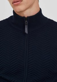 Mornarica zip-up dukserica s visokim ovratnikom i teksturiranim ribljim kostima, s crnim patentnim zatvaračem i oznakom za vješanje.