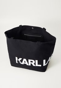 Sac fourre-tout noir en tissu durable. Présente un grand logo blanc, deux poignées et une poche zippée à l'intérieur pour un rangement sécurisé.