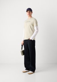 Beige bavlněné tričko s jemným potiskem, přetažené přes bílé dlouhé rukávy. Tmavé wide-leg kalhoty, bílé tenisky a zelená kabelka.