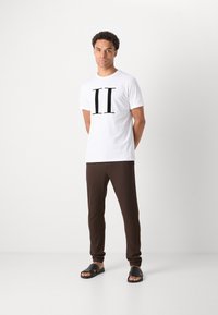 Camiseta blanca con un gran estampado negro de "II", combinada con pantalones marrones ajustados y sandalias negras. Diseño sencillo y corte casual.