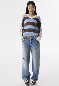 Pull tricoté rayé en marron, bleu et crème ; associé à un jean baggy déchiré bleu clair et des baskets multicolores.
