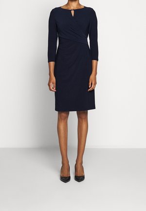 Frau trägt ein knielanges, langärmeliges navyblaues Kleid mit kleinem Ausschnitt vorne und schwarzen spitzen Schuhen vor neutralem Hintergrund.