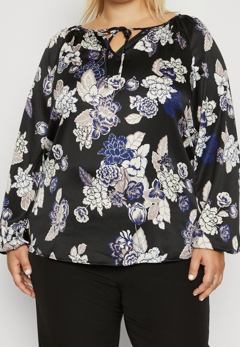Blouse noire à manches longues avec motifs floraux bleus, blancs et beiges, dotée d'une encolure échancrée avec un détail noué, portée avec un pantalon noir.