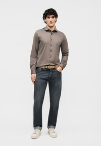 Man standing with one hand in pocket wearing taupe button-up shirt, dark jeans, tan belt, and white sneakers against plain background.