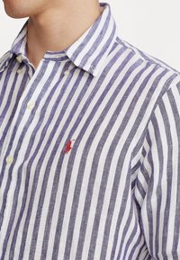 Herrskjorta med knappar i vit och marinblå randig design, med en liten röd logotyp på bröstet, klassisk krage och textilstruktur.