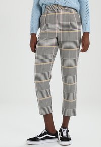 Pantaloni a quadri bianco e nero con accenti gialli, taglio corto, design sartoriale e texture liscia, abbinati a sneakers nere.