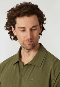 Olivgrön polo skjorta i mjukt tyg; har en krage, korta ärmar och en diskret logotyp på vänster sida.