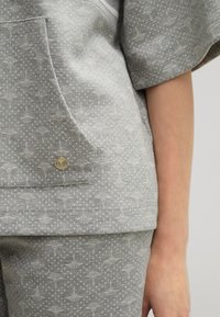 Grau gemusterte Bluse mit gestickten Designs, die eine Tasche und subtile Punkte aufweist. Weicher Stoff mit leicht strukturierter Oberfläche.