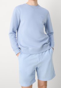 Felpa a girocollo di colore azzurro chiaro realizzata in tessuto morbido, con un design semplice e polsini a costine, abbinata a pantaloni corti azzurri chiari.