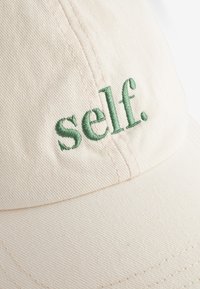 Pet met een lichtbeige katoenen twill met een gebogen klep, met groene geborduurde tekst "self." op de voorkant. Getextureerde stof.