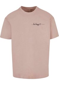 Kortærmet t-shirt i lyserød bomuld med rund halsudskæring og trykt sort tekst "vær glad" med et hjerteakcent på brystet.