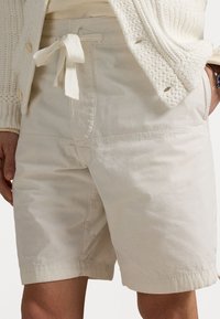 Bežové bavlněné šortky s nastavitelným páskem, vybavené předními kapsami a hladkým, mírně strukturovaným materiálem.