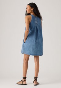 Denim dress with a loose fit, sleeveless design, blue color, and back pleat detail. Features side pockets and a hemmed finish.