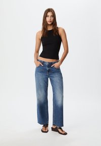 Svart ärmlös topp, ljusblå vida jeans med blekta kanter och bruna flip-flops, händer i fickorna, neutral bakgrund.