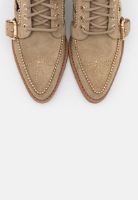 Chaussures en daim beige à bout pointu avec lacets, brides à boucle et clous dorés décoratifs formant des motifs sur la zone des orteils.