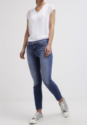 Frau trägt weiße kurzärmelige Bluse, blaue Skinny-Jeans und silberne glitzernde Sneaker, steht mit einem Bein leicht nach vorne.