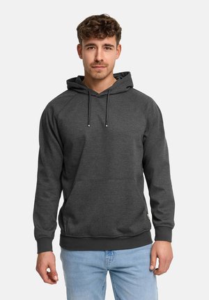 Mężczyzna noszący ciemnoszarą bluzę z kapturem i kieszenią z przodu oraz niebieskie dżinsy, stojący na tle jednolitego jasnego tła.