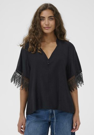 Melns tops ar V veida kakla izgriezumu, pogas detaļu un īsām piedurknēm ar smalku melnu mežģīņu apdari. Audums šķiet viegls un strukturēts.