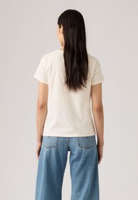 T-shirt en coton à manches courtes de couleur crème clair, associé à un jean en denim bleu à coupe ample avec poches latérales et taille haute.