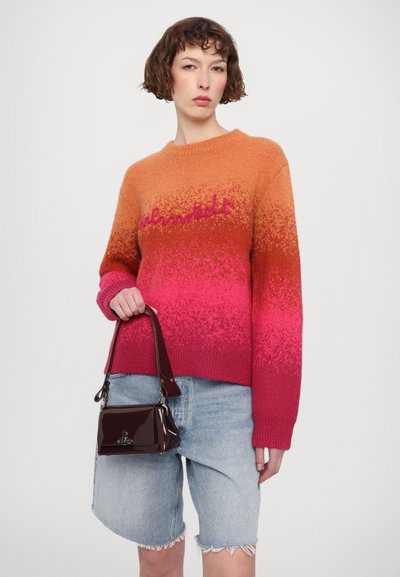 Sweter z gradientowym efektem ombre od pomarańczowego do różowego, z wyraźną teksturą dzianiny i szczegółowym napisem. Modelka trzyma bordową torebkę na ramię z lakierowanej skóry.