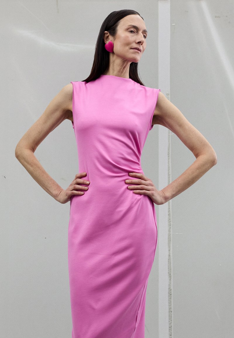 Přiléhavé šaty bez rukávů v zářivě růžové látce, s hladkou texturou a jemným řasením, stylizované s výraznými růžovými náušnicemi.