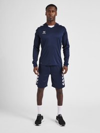 Marineblauwe sportieve longsleeve shirt met witte accenten en logo, gecombineerd met bijpassende shorts met witte chevronstrepen. Zwarte sportschoenen.