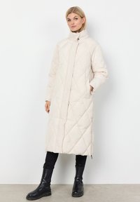 Lange, lichtbeige gewatteerde jas met een hoge kraag, voorrits en zijzakken, gecombineerd met zwarte, aansluitende broeken en zwarte laarzen tot de knie.