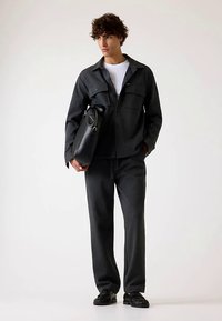 Dunkelgraue Männerjacke mit Knöpfen und zwei Brusttaschen, kombiniert mit passenden Hosen und einer schwarzen Tasche. Der Stoff hat eine glatte Textur.