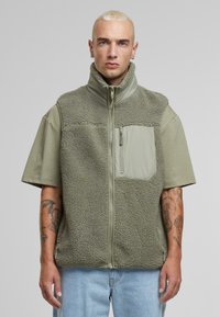 Fleece vest in olive green with a high collar, front zip closure, and a textured body. Features a smooth chest pocket with zipper.