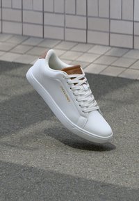 Weiße Low-Top-Sneaker mit braunen Akzenten und "Jack & Jones"-Logo, schwebend über grauem Pflaster neben einer gefliesten Wand.