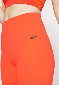 Gros plan sur le ventre portant un short de sport taille haute orange vif et un soutien-gorge de sport assorti, avec le logo noir "4F" sur le short.