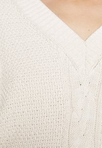 Gros plan sur un pull à col en V tricoté de couleur crème, présentant des motifs texturés sur une peau claire.