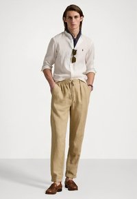 Μπεζ λινά παντελόνια με ελαστική μέση, λευκό λινό πουκάμισο και καφέ μοκασίνια. Τα αξεσουάρ περιλαμβάνουν γυαλιά ηλίου και μαντήλι με σχέδιο.