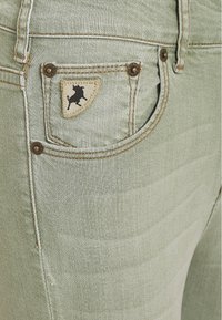 Světle zelené džíny z denimu se šitou kapsou s hnědou nášivkou a černým logem býka. Označeny měděnými kovovými detaily.