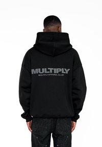 Felpa nera con grande scritta grigia "MULTIPLY" sulla schiena. Caratterizzata da una tasca a canguro frontale e cappuccio con cordino. Il tessuto sembra morbido e strutturato.