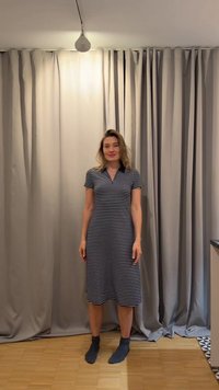 Γυναίκα στέκεται σε εσωτερικό χώρο μπροστά σε γκρίζες κουρτίνες, φοράει μαύρο-λευκό ριγέ φόρεμα και σκούρες κάλτσες μέχρι τον αστράγαλο σε ξύλινο πάτωμα.