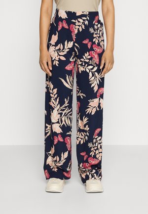 Pantalon large marine avec imprimé papillons roses et rouges et motifs floraux, porté avec un haut beige et des baskets blanches.