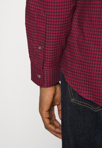 Rood en zwart geblokt overhemd met lange mouwen, voorzien van grijze knopen en een gebogen zoom, gecombineerd met donkere spijkerbroeken.