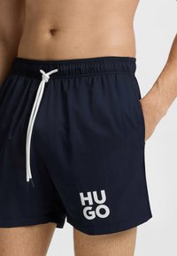Marina simbyxor med elastisk midja och vit dragsko. Har en iögonfallande "HUGO"-logotyp i vitt på vänster ben. Lättviktig tyg.