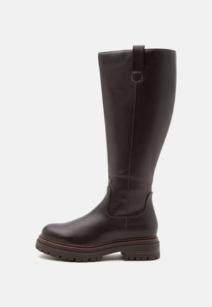 Botte haute en cuir marron foncé avec semelle épaisse et languette latérale sur fond uni gris clair.