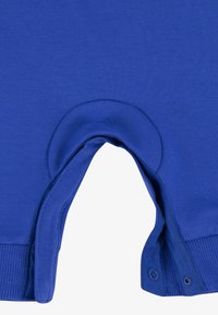 Macacão de algodão azul com uma abertura redonda, punhos largos em nervura e fechos de botão de pressão para vestir facilmente. Textura suave com cor sólida.