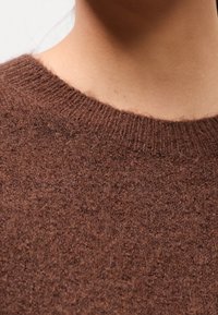 Pull en maille marron avec une texture apparente, un col rond et des fibres douces, mettant en avant chaleur et confort.