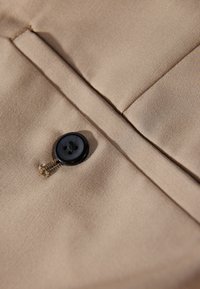 Beiger Stoff mit horizontaler Textur, auf dem ein schwarzer Knopf auf einer Klappe angenäht ist. Die Nähte sind entlang der Kanten sichtbar.